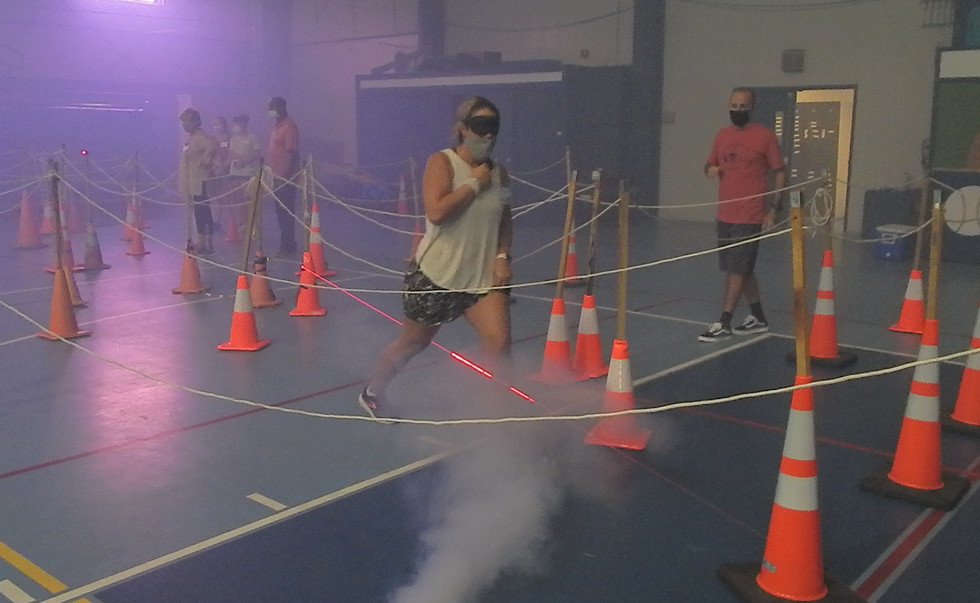 A woman in a blindfold and mask makes her way through a rope maze. It is held up by stakes stuck into traffic cones. She is stepping over a laser beam obstacle and there is fog all around. A man stands nearby giving her directions.