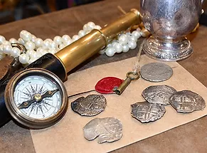 Authentic pirate coins rest on a sealed envelope on a table with an old-fashioned key, a compass, a telescope, a string of pearls, and a silver tankard