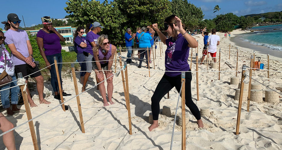 A blindfolded woman in a purple shirt with her hands up in the air moves through a stake and rope maze on the beach. Another woman in a purple shirt stands nearby giving her directions. Several other people in purple shirts stand and watch.