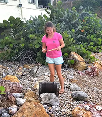 Woman in pink shirt and blue shorts stands on a rocky beach by an open treasure chest reading a piece of paper.