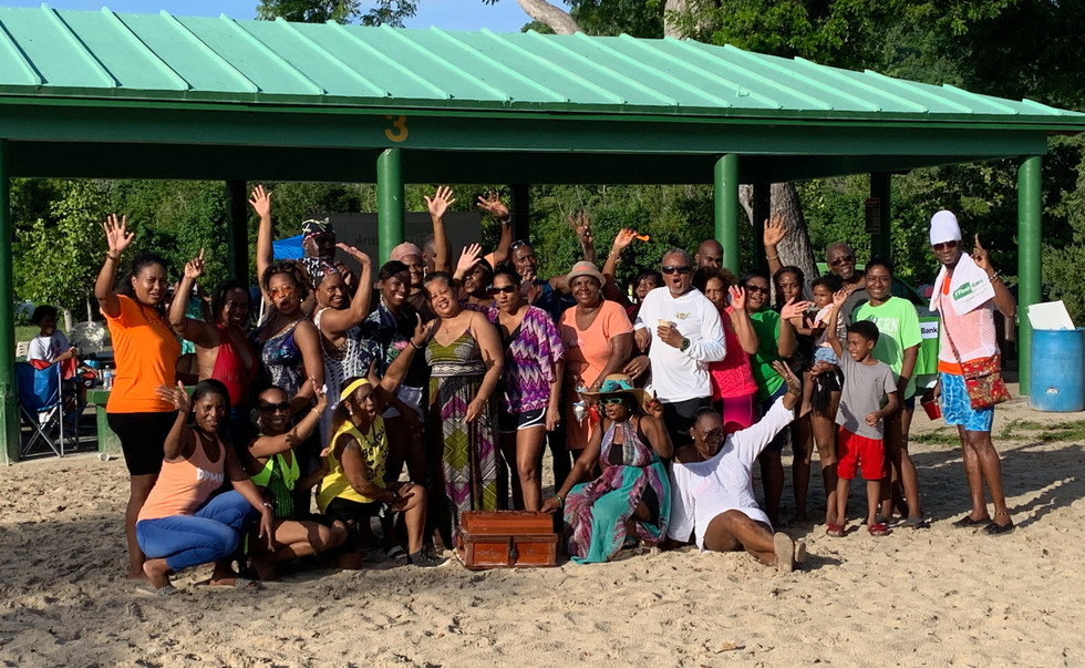 A large group of people stand in front of a covered pavilion on the beach. There is a treasure chest in front of them.