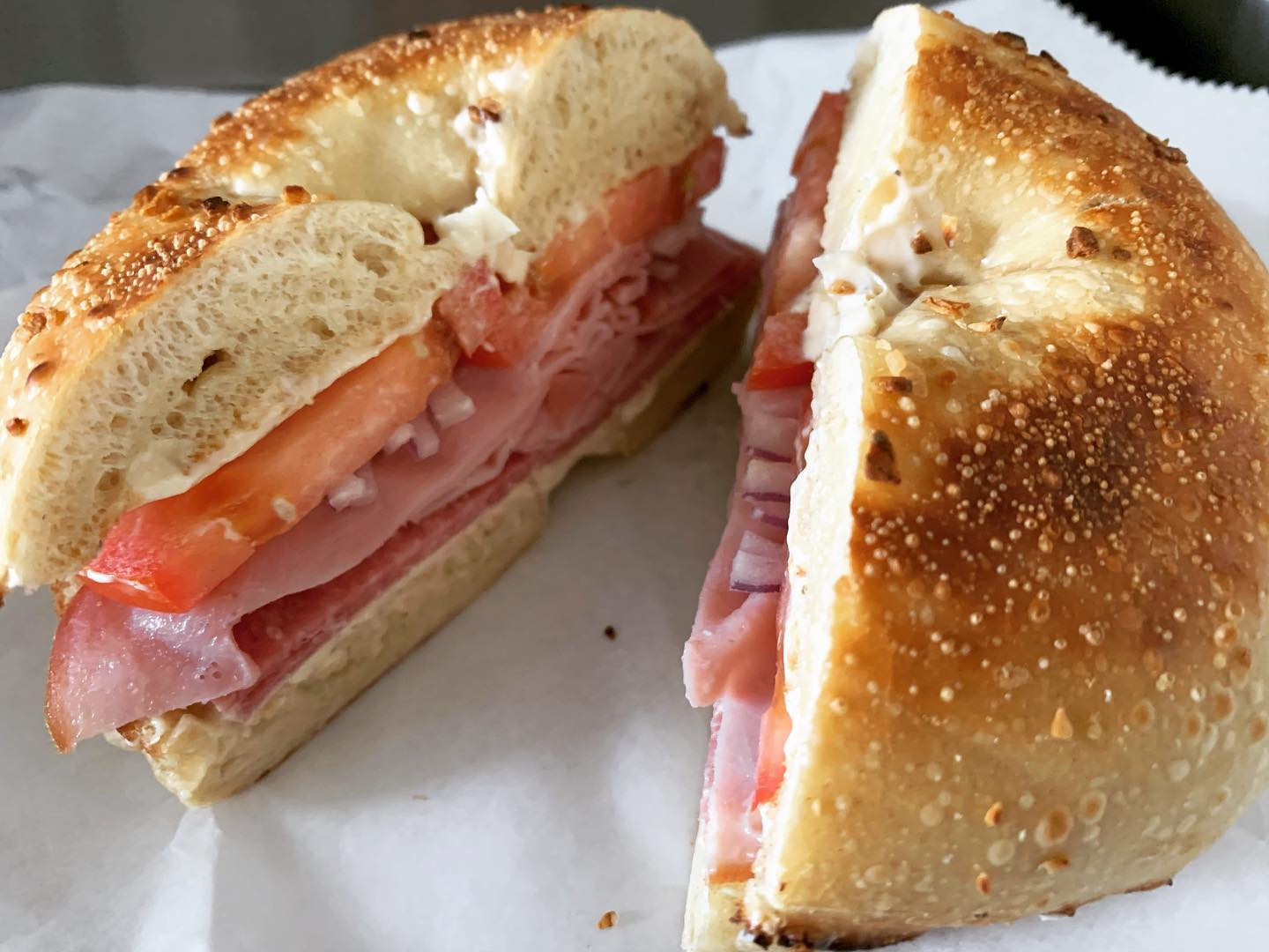 A bagel sandwich with cream cheese, tomatoes, and cold cuts cut in two sits on white parchment paper.