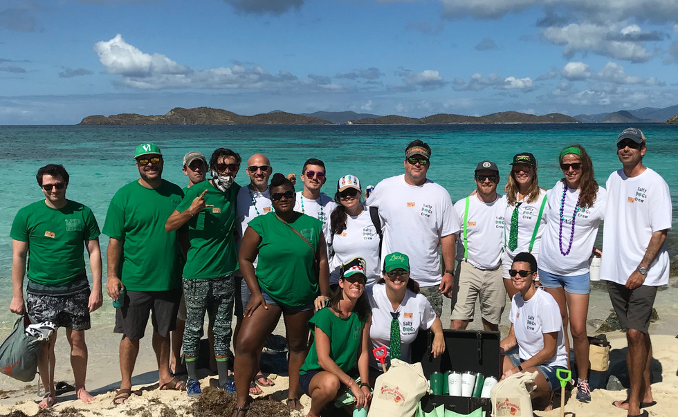A large group of people pose on the beach in front of the ocean. About half the group is in green t shirts while the other half is in white. They are posing with two canvas bags with the Tropical Treasure Hunt logo and a treasure chest full of green and white drink tumblers.