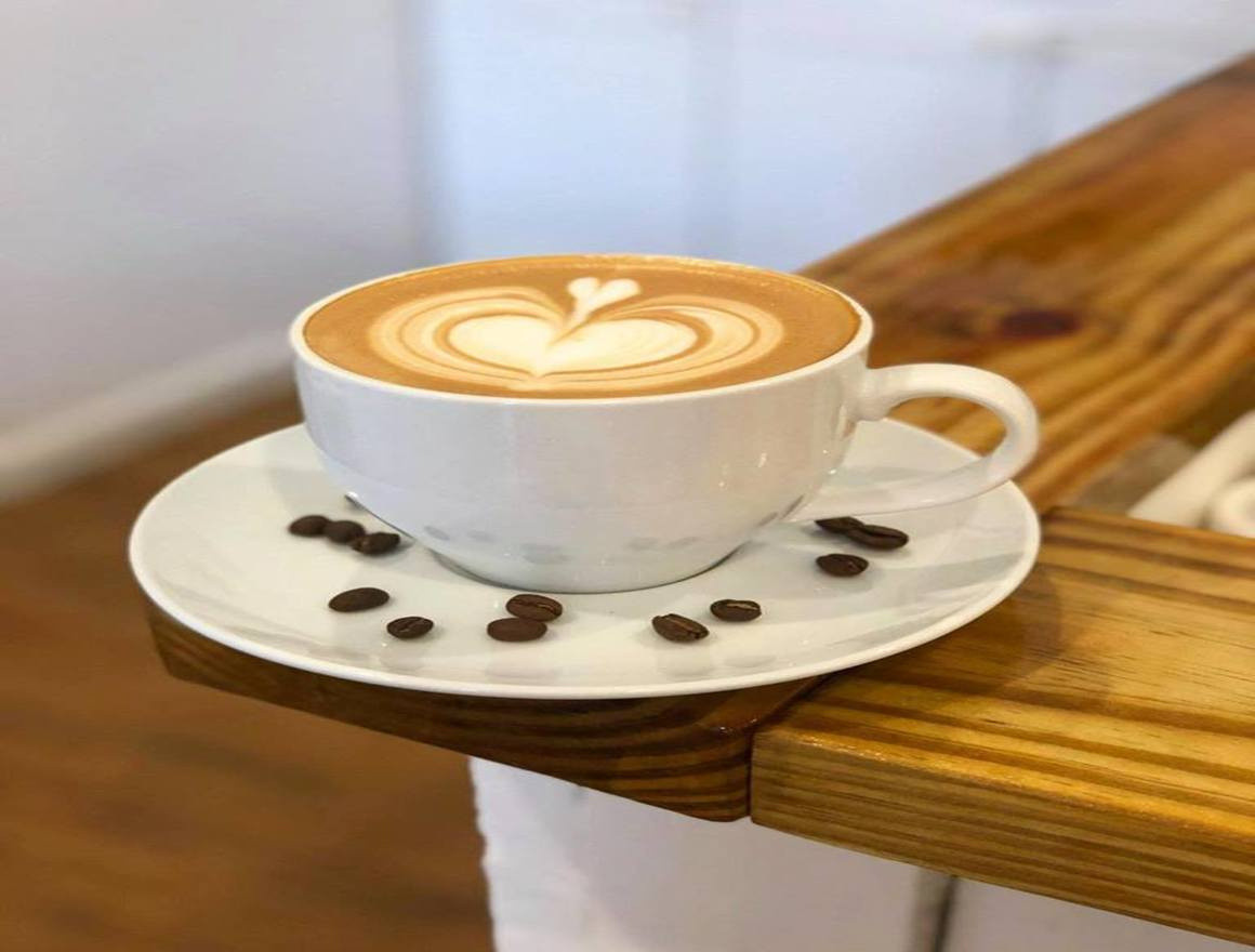 Cup of coffee with a heart design in the froth sits on a saucer with some coffee beans on it on the edge of a counter