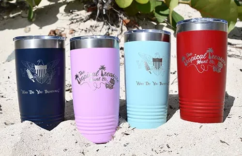 Tumblers in navy, lavender, light blue, and red featuring Tropical Treasure Hunt logo and VI crest sit in the sand