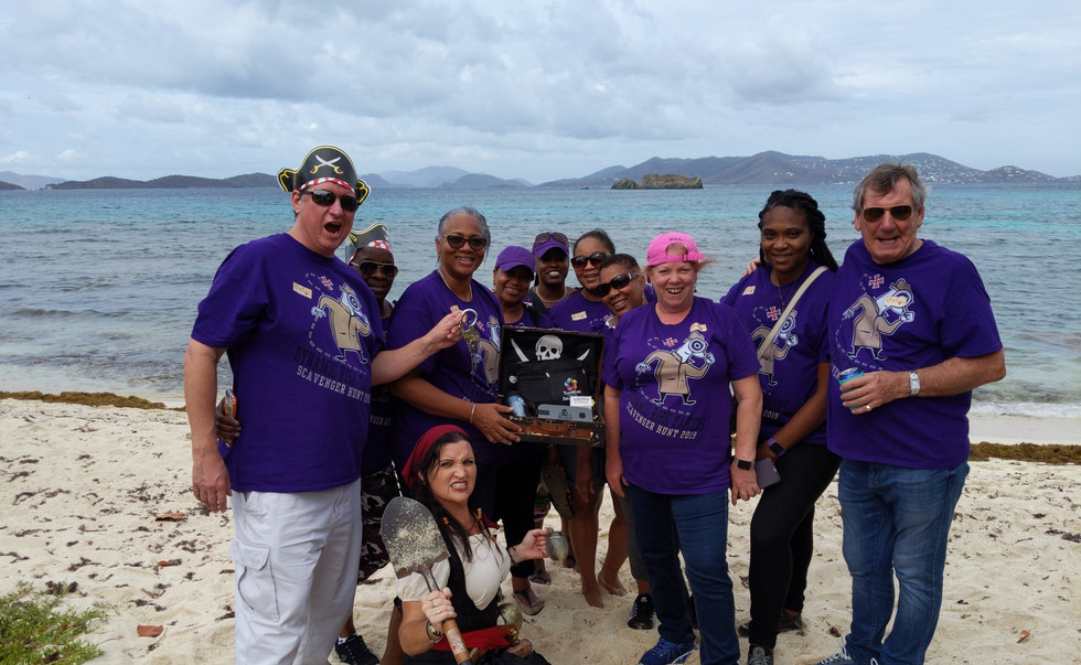 Ten people in purple shirts pose with a treasure chest on a beach in front of the ocean. A woman dressed as a pirate holding a metal tankard and a shovel crouches in front of them.