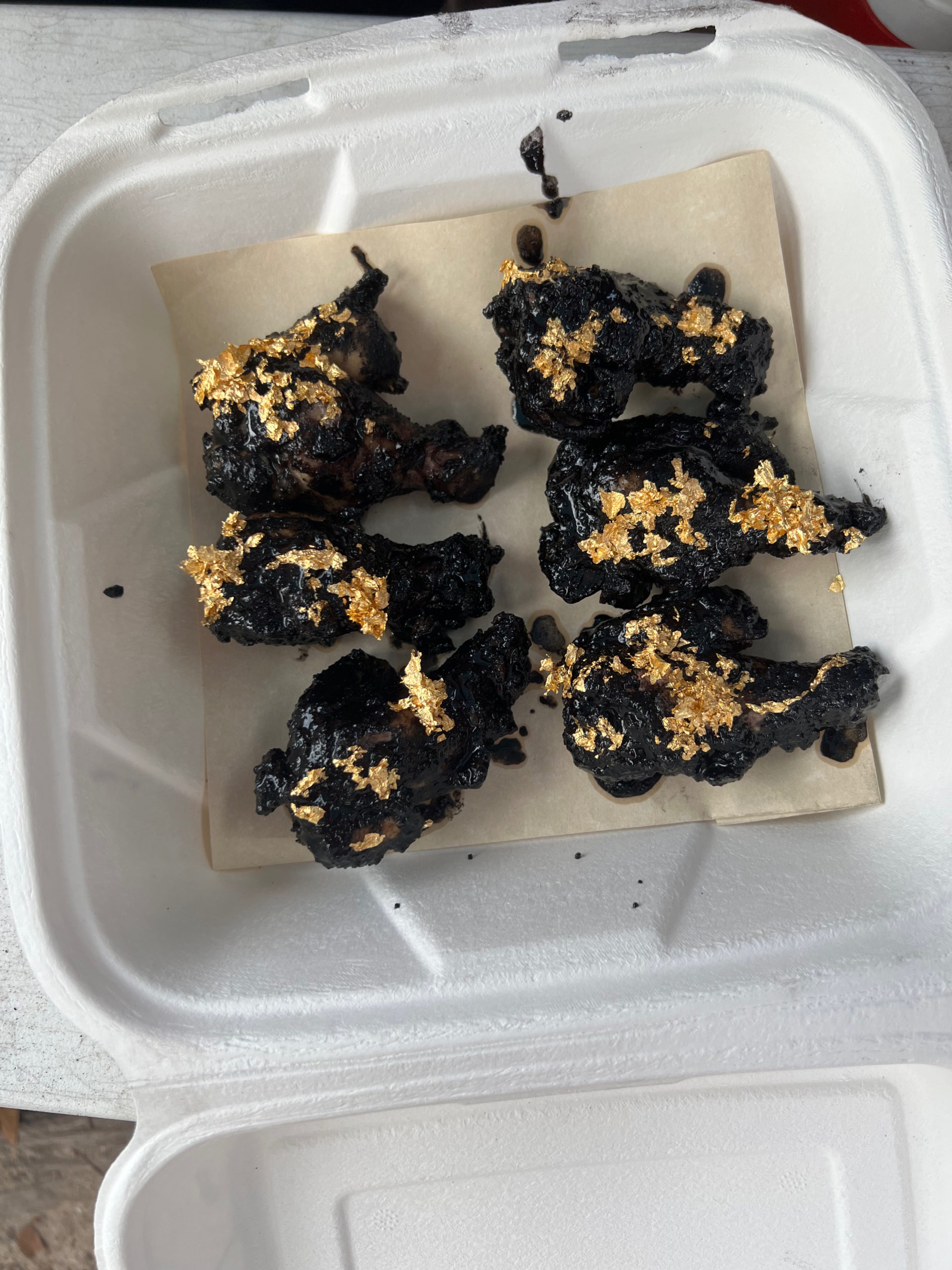 6 black chicken wings with gold flake rest on a paper in a cardboard take out container.