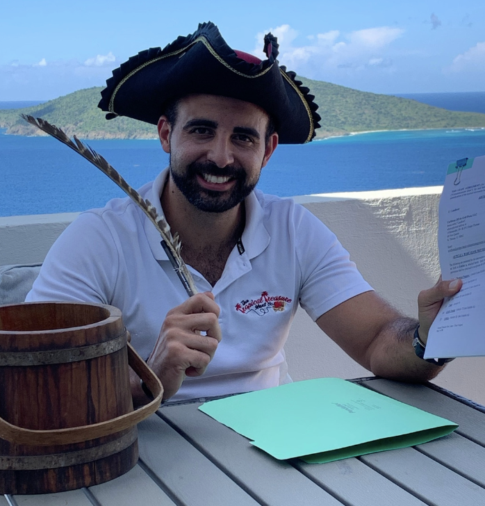 Man in a three-corner hat sits at a table with a view of the ocean and an island behind him. He holds a quill pen. A bucket and a folder rest on the table in front of him.