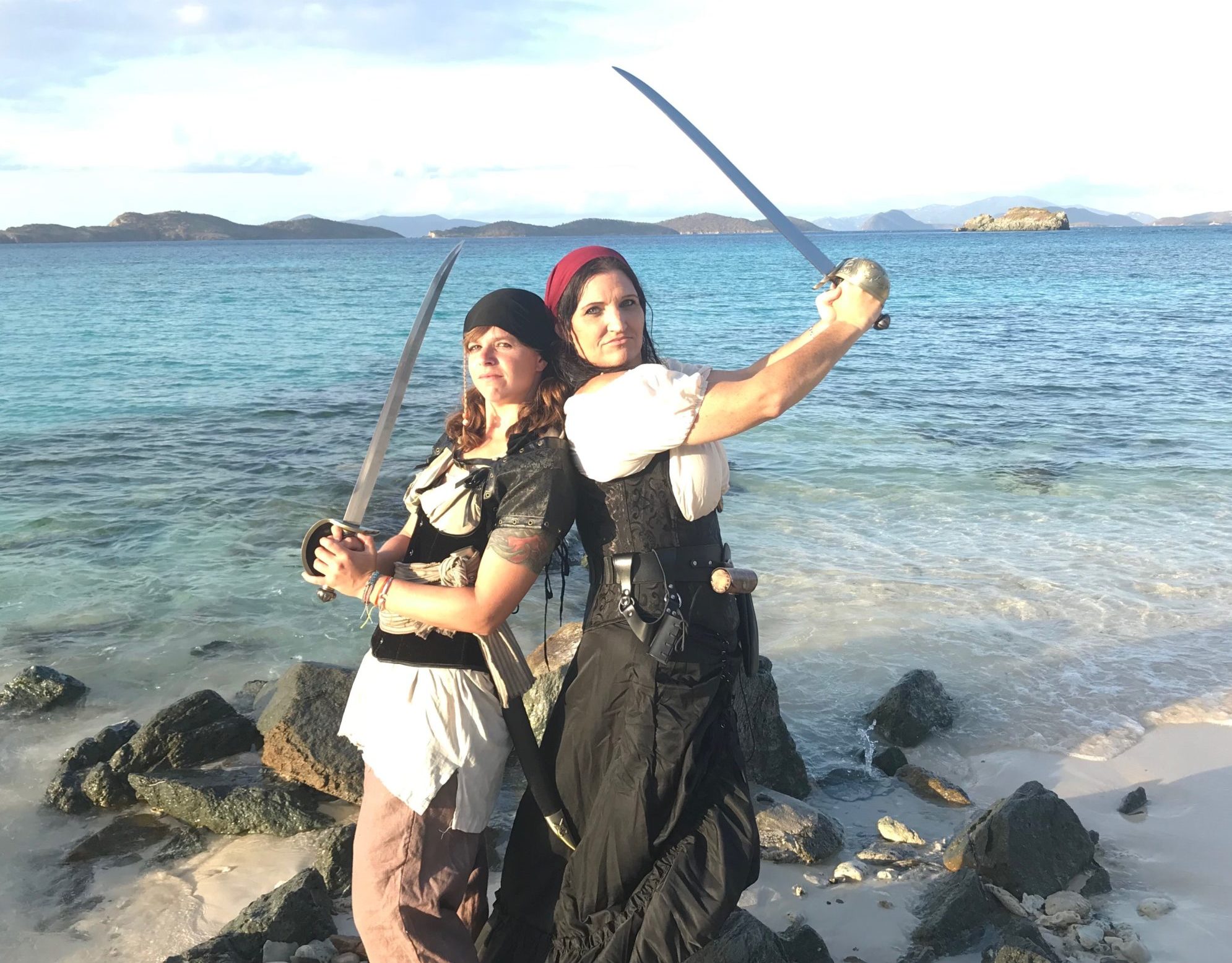 Two pirates pose back to back on a rocky beach, brandishing swords