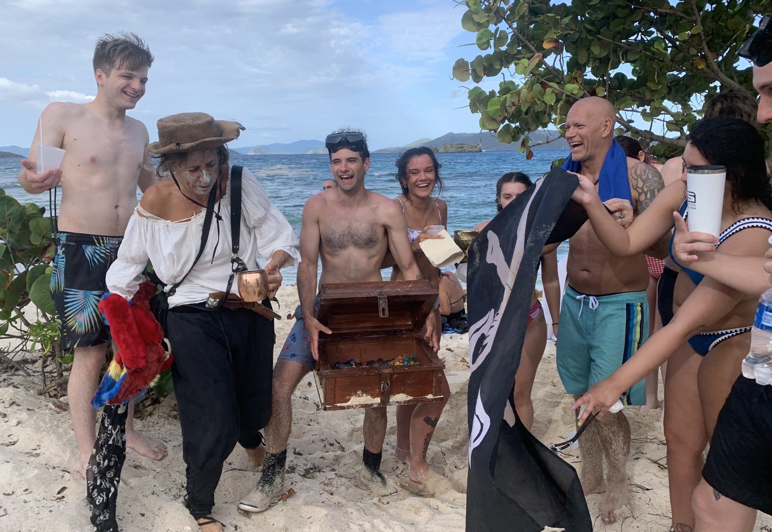 A group poses on a beach with a treasure chest, a Jolly Roger, and Anne Bonny.
