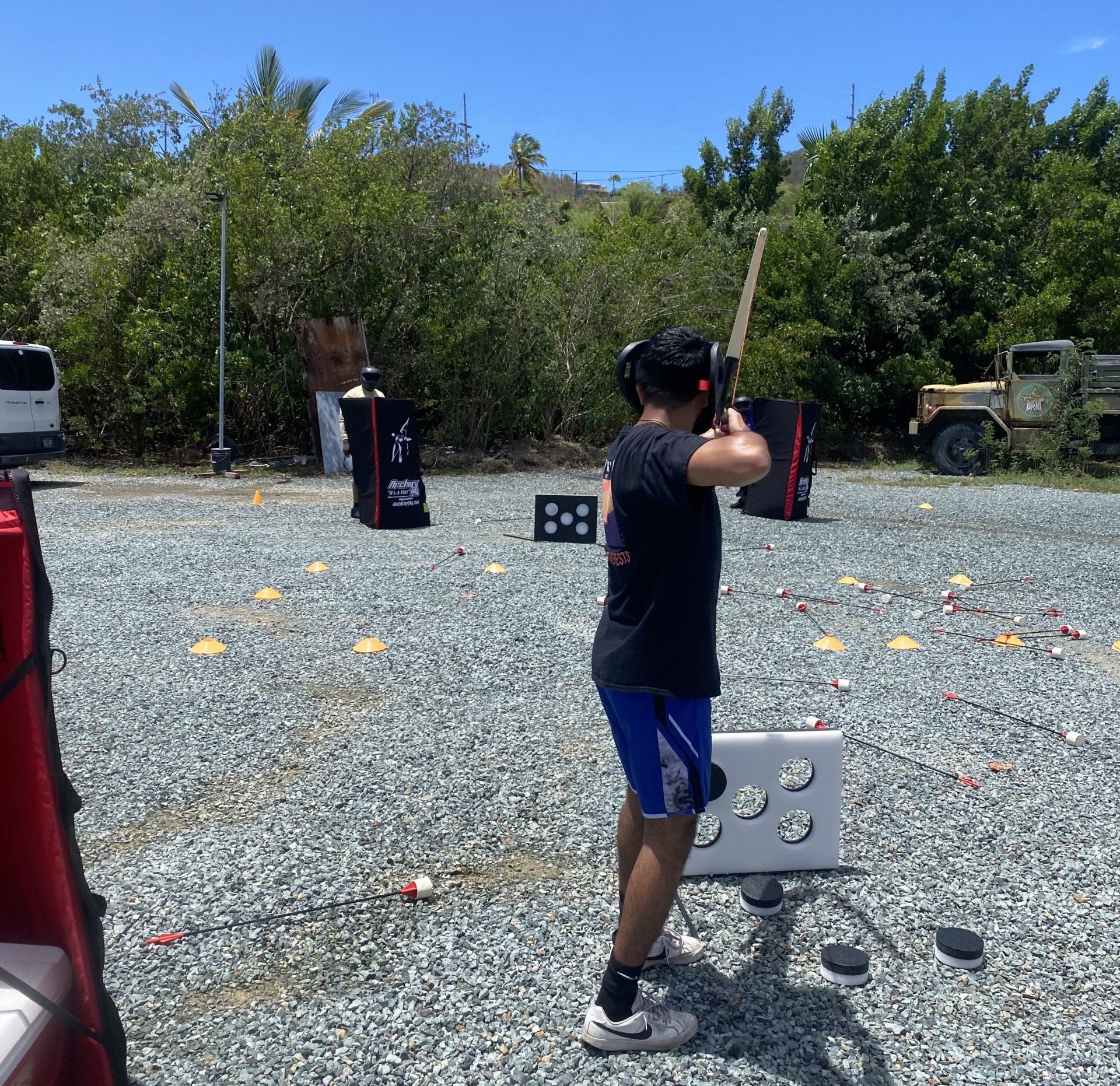 A man in a gravel parking lot and a protective face mask draws a bow and arrow toward a target