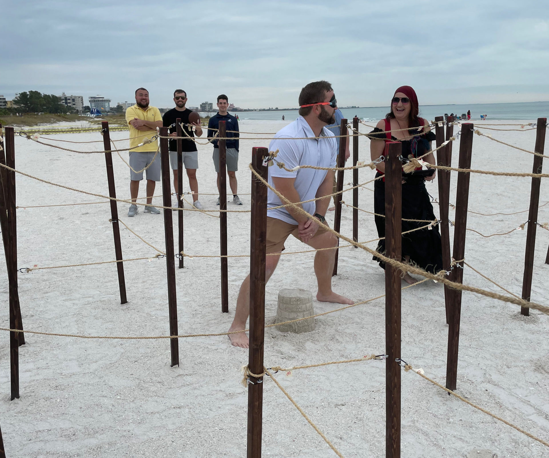 A blindfolded man walking through a rope maze takes a big step over a sandcastle. A woman in a pirate costume with an iPad for scorekeeping looks on.