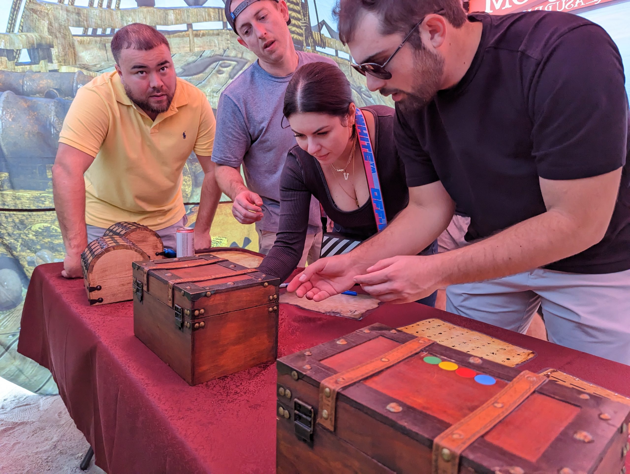 Four people lean over a table filled with treasure chests and paper puzzles.