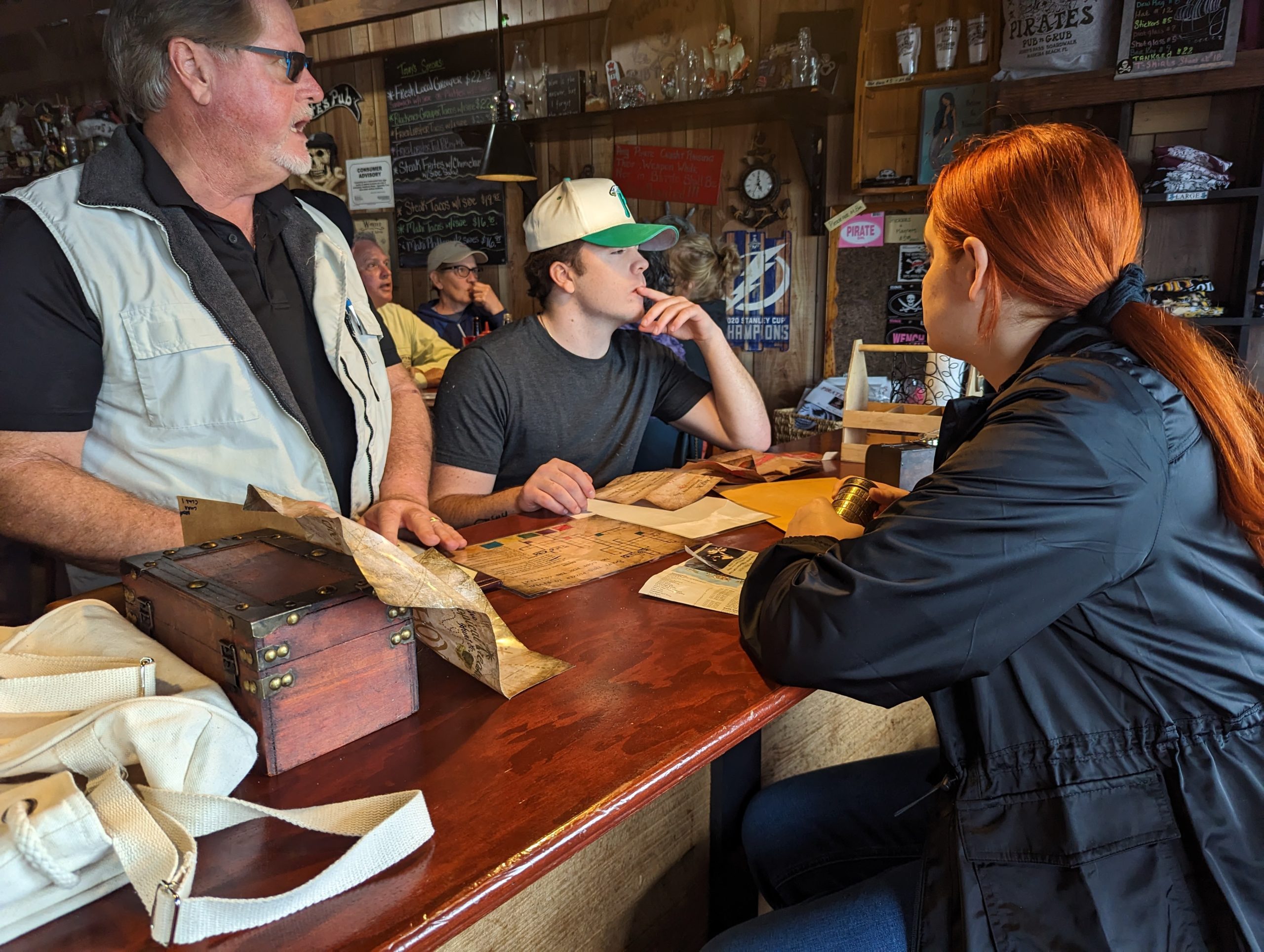 Three people sit at a bar with clues and puzzles before them.