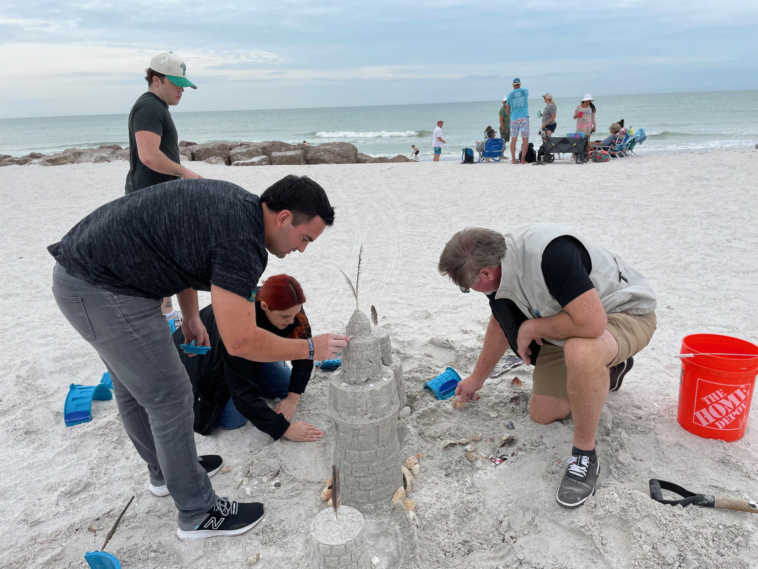 Three people bend over a sandcastle on the beach. There are sandcastle molds, shells, and seaweed in the castle.