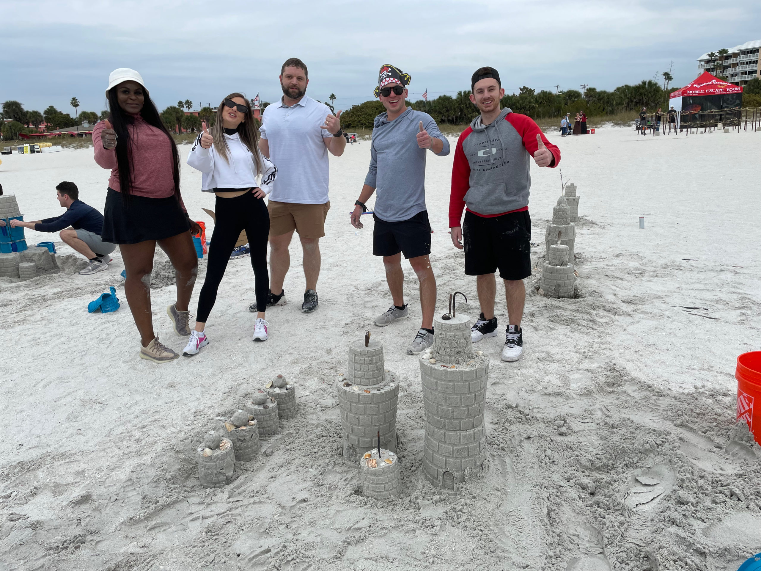 Four people with thumbs up stand behind a sandcastle with two large tiers and several small tiers on the beach.