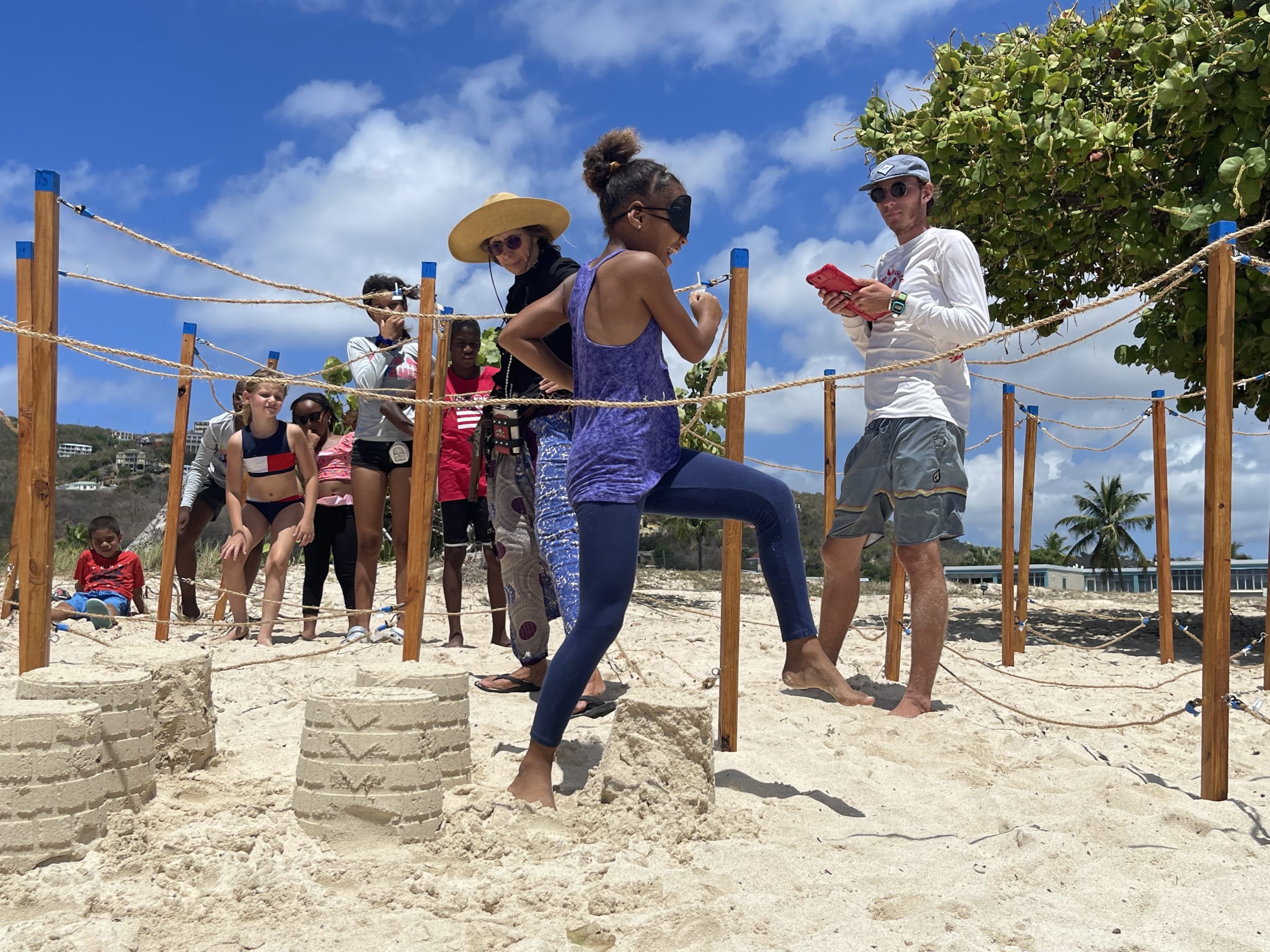 A blindfolded girl steps over a sandcastle while going through a maze made of stakes and ropes. A man with a baseball hat an an iPad stands near by. A woman with a sun hat and a group of children are also nearby watching.