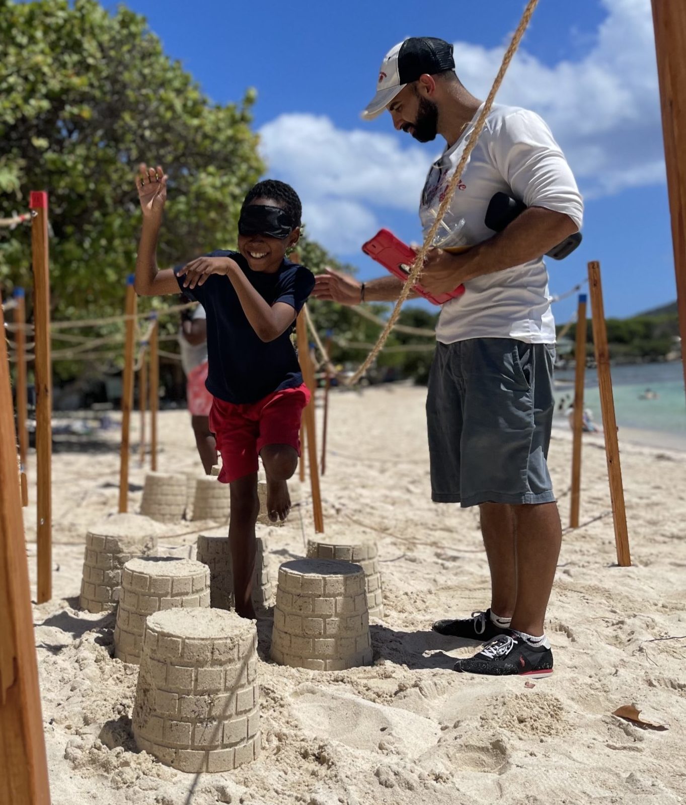 A blindfolded boy steps over a sandcastle while walking through a maze made of poles and ropes. A man with a baseball cap and an iPad stands nearby.