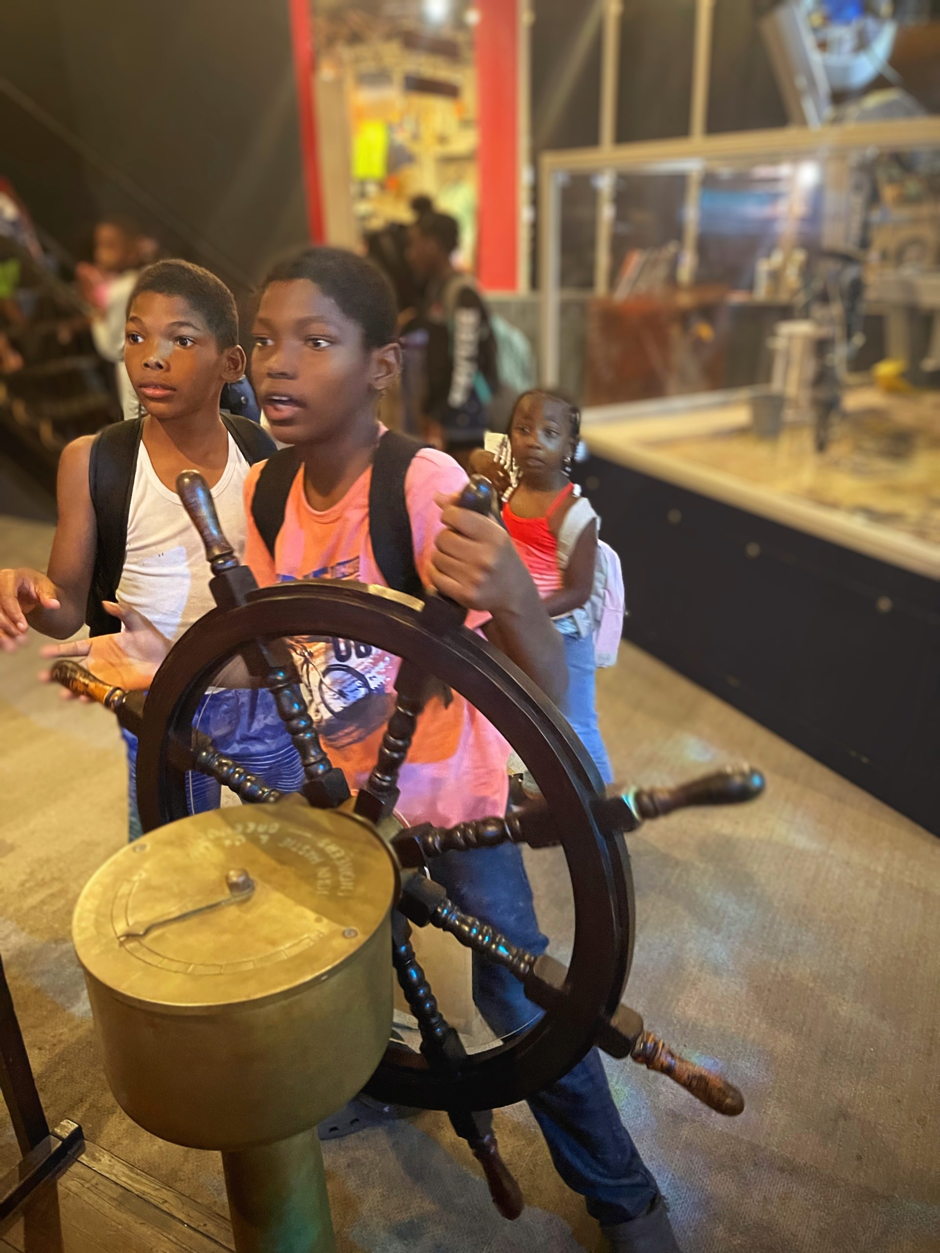 Two boys spin a ships wheel while staring intently at something behind it (out of the frame).