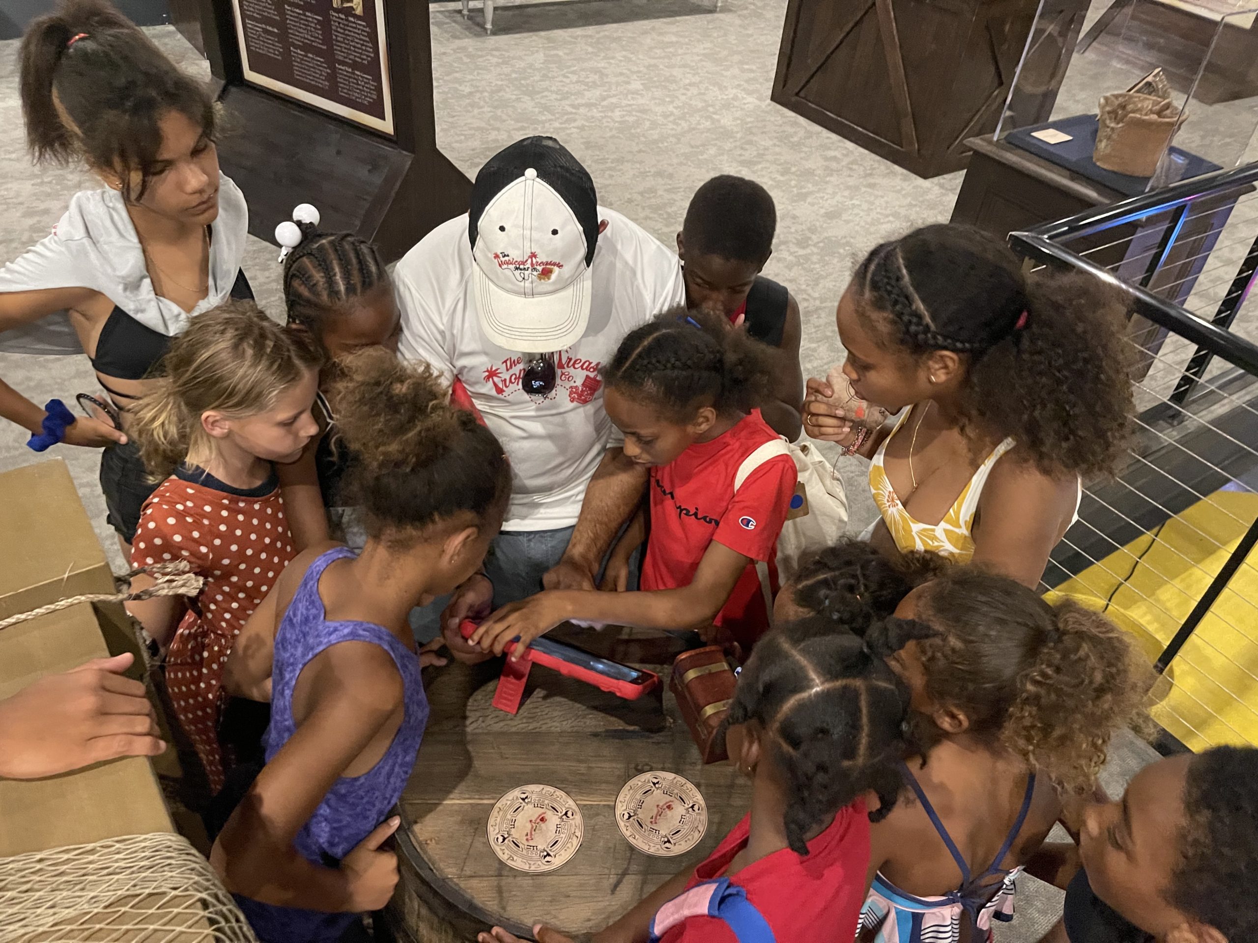 A group of children stands around a barrel with an iPad on top. A man stands in the midst helping the children use the iPad.