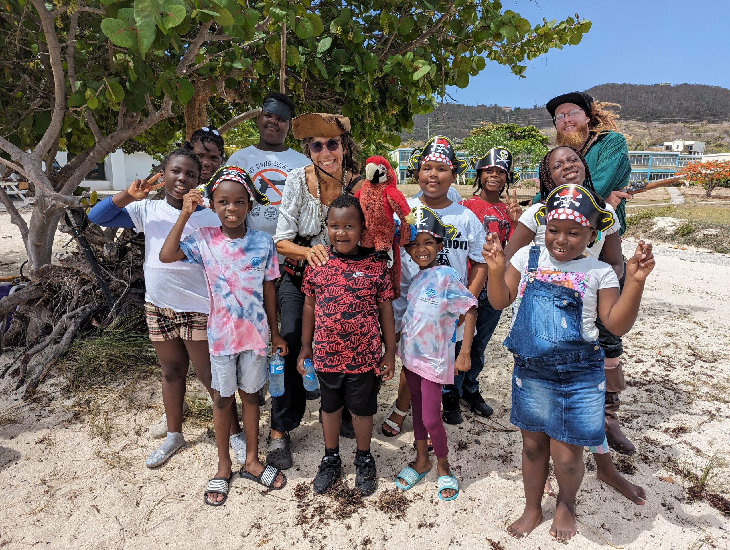A group of children wearing paper pirate hats pose on a beach next to a mangrove tree with two adults dressed as pirates.