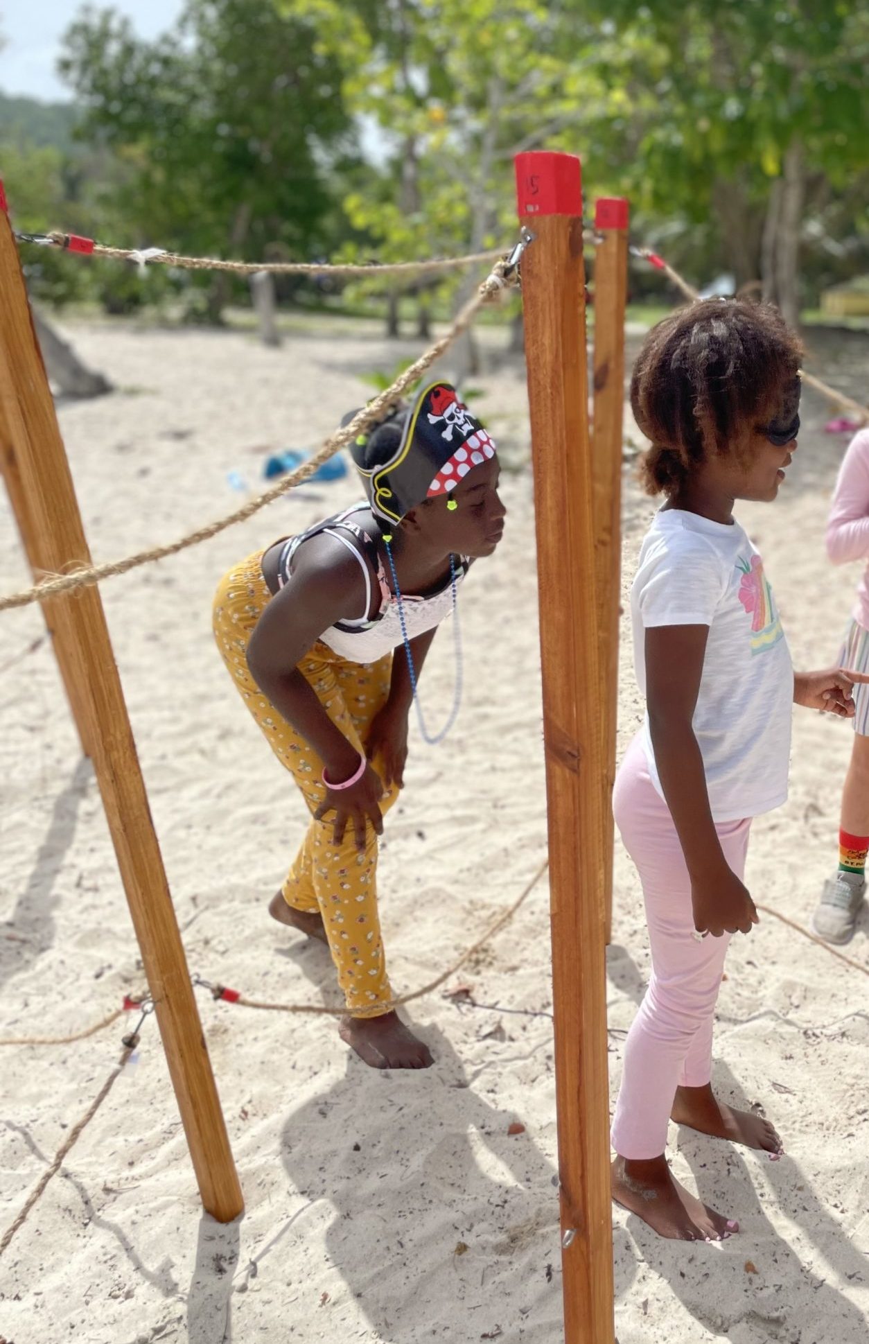 A girl with a blindfold stands in a maze made of stakes and ropes. Another girl with a paper pirate hat leans toward her, giving directions.