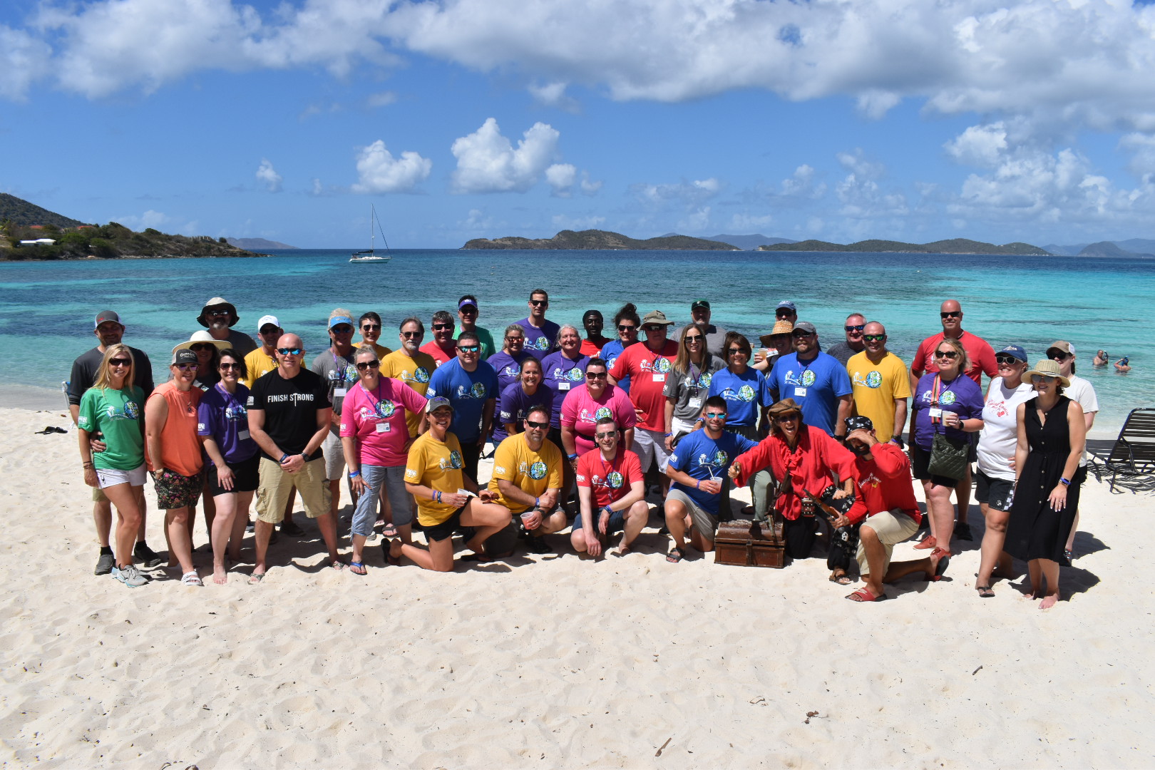 A group of about 60 people in different colored shirts pose on a beach. There is a treasure chest and a woman dressed as a pirate in the middle of their group.