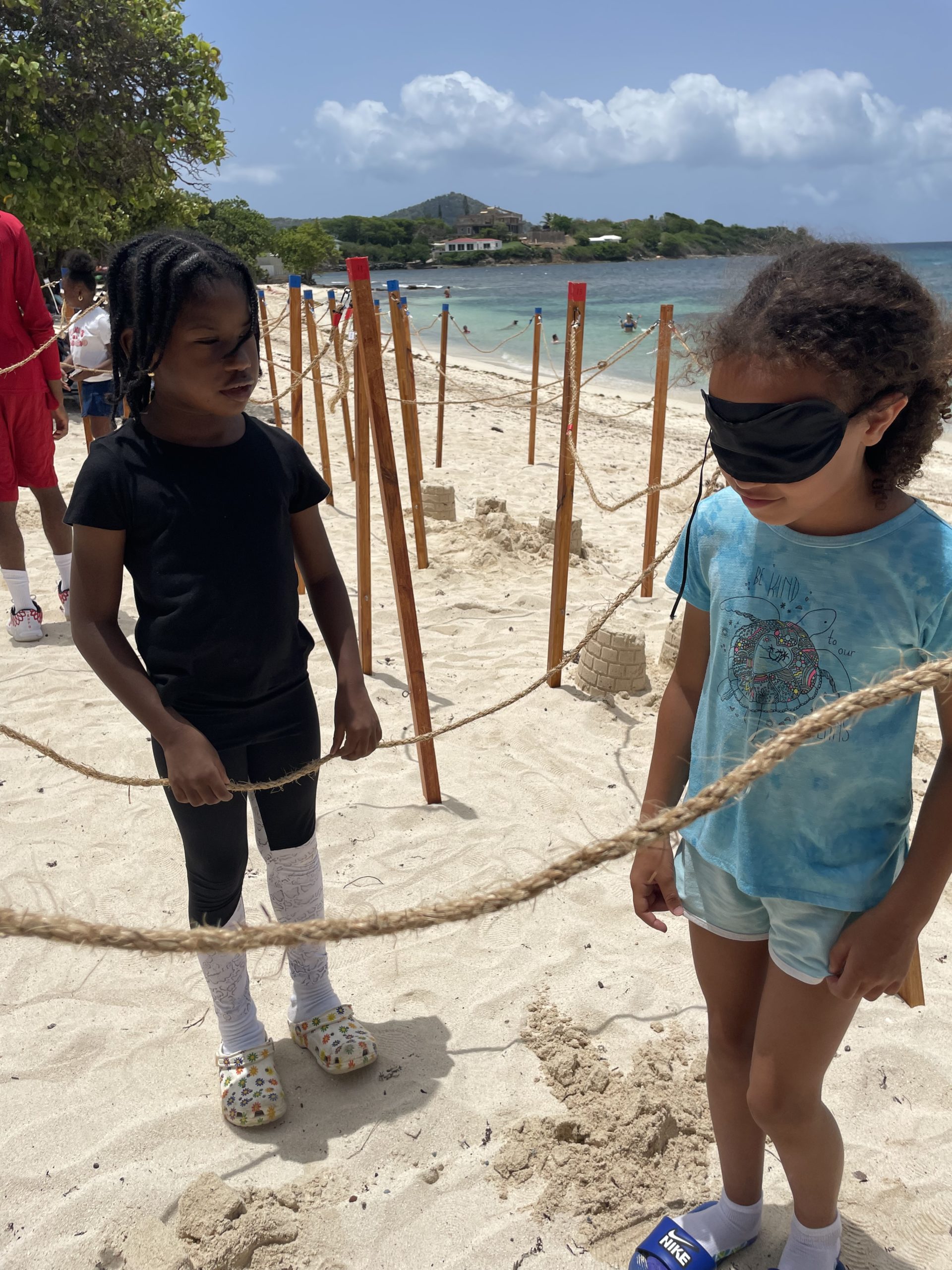 One child in slides and a blue tshirt and shorts wears a blindfold in a rope maze. Another child wearing Crocs and all black stands nearby.